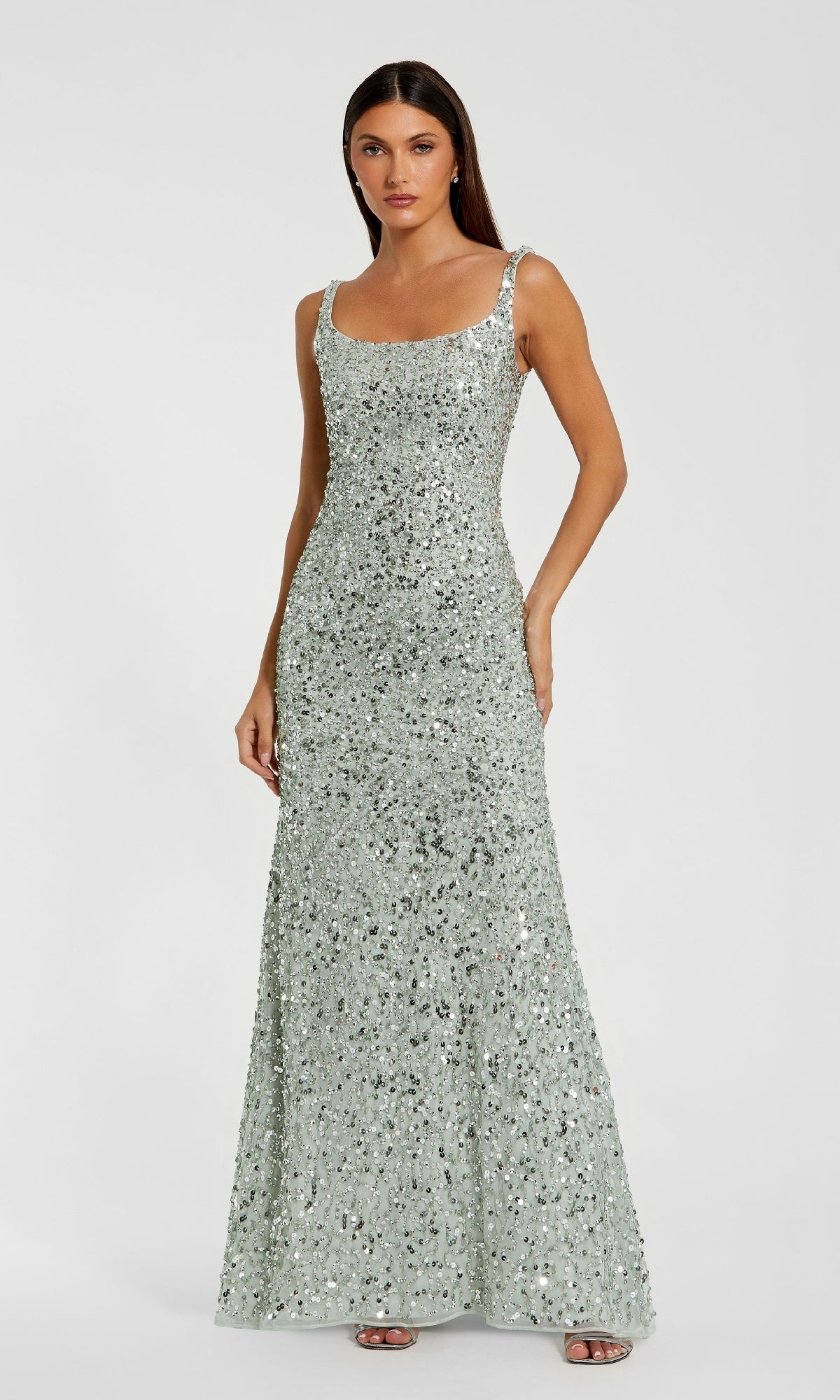 A woman wears the Sparkly Long Sequin Formal Dress: Mac Duggal 10705, a floor-length, silver, open-back gown with a scoop neckline and fitted silhouette, standing against a plain light background.